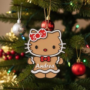 May include: A Christmas tree ornament featuring a cartoon cat design with a red bow and the name "Andrea". The ornament is brown, white, and red, and is hanging on a Christmas tree with other ornaments.
