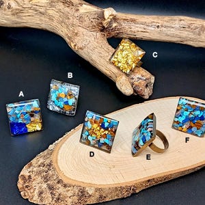 Peut inclure: Six bagues carrées avec divers incrustations colorées de pierres et de feuilles d'or. Les bagues sont exposées sur une tranche de bois et un morceau de bois flotté sur fond noir. Les bagues ont des bandes de couleur bronze.