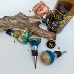 Può includere: Una collezione di tappi decorativi per bottiglie con design unici. Uno presenta un cristallo rosa e bianco, un altro una casa in miniatura e altri elementi a tema spiaggia. I tappi sono realizzati in resina e metallo.