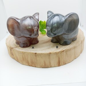 Peut inclure: Deux petites figurines d'éléphants, une marron et une grise, sont posées sur une tranche de bois. Les éléphants ont de grandes oreilles et des trompes, avec une finition brillante. L'éléphant marron est étiqueté "A" et l'éléphant gris est étiqueté "B".