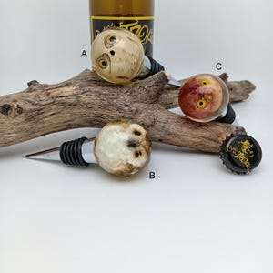 Puede incluir: Tres tapones decorativos para botellas con diseños únicos. Uno tiene una cara de búho, otro tiene una apariencia veteada y el tercero tiene un patrón rojo y amarillo. También se ven una botella y un tapón de botella.