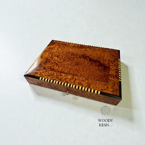 Puede incluir: Una caja de madera rectangular con un acabado brillante. La caja presenta un cálido patrón de madera de raíz marrón en la parte superior y los lados, con detalles en negro y dorado. La caja tiene un pequeño cierre de metal. El logotipo "WOODY KESH LLC" es visible.