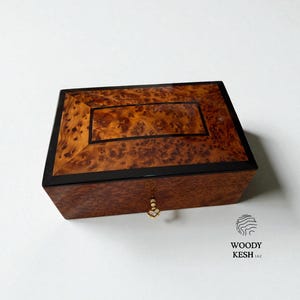 Puede incluir: Una caja rectangular de madera con tapa de madera de raíz y borde negro. La caja tiene un ojo de cerradura dorado y un diseño decorativo en la tapa. La caja probablemente se utiliza para guardar joyas u objetos pequeños.