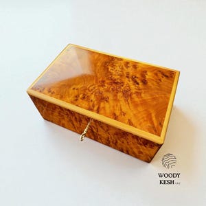 May include: A rectangular wooden box with a glossy finish. The box is a warm, golden brown color with a unique wood grain pattern. A small, gold-colored latch is visible on the front. The logo "WOODY KESH LLC" is in the bottom right corner.