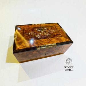 May include: A rectangular wooden box with a glossy finish. The lid features a decorative inlay with floral and geometric patterns. Gold accents and a keyhole are visible. The box is likely for jewelry or keepsakes.