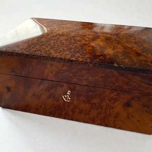 May include: A rectangular wooden box with a glossy finish. The box is made of a dark brown wood with a unique, swirling grain pattern. It features a gold-colored clasp and a slightly angled top.