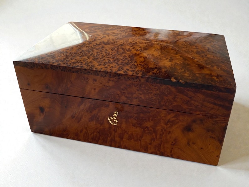 May include: A rectangular wooden box with a glossy finish. The box is made of a dark brown wood with a unique, swirling grain pattern. It features a gold-colored clasp and a slightly angled top.