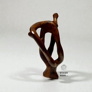 May include: A brown wooden sculpture of an abstract human form. The sculpture has a smooth, polished finish and is in a contorted pose, with limbs intertwined. The sculpture is set against a white background. The text "WOODY KESH.UK" is visible.