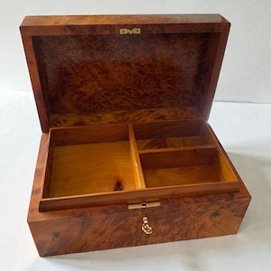 May include: Open wooden box with a hinged lid, featuring a rich, patterned burl wood exterior. The interior is divided into compartments. The box is open, revealing the interior. The box has a brass-colored clasp and hinges. The box is likely for jewelry or keepsakes.