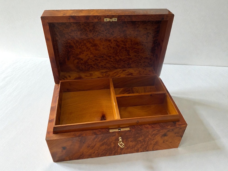 May include: Open wooden box with a hinged lid, featuring a rich, patterned burl wood exterior. The interior is divided into compartments. The box is open, revealing the interior. The box has a brass-colored clasp and hinges. The box is likely for jewelry or keepsakes.