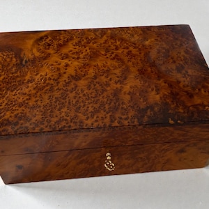 May include: A rectangular wooden box with a rich, warm brown burl wood grain finish. The box has a small, ornate gold-colored keyhole and a matching pull. The wood grain features a unique, swirling pattern.