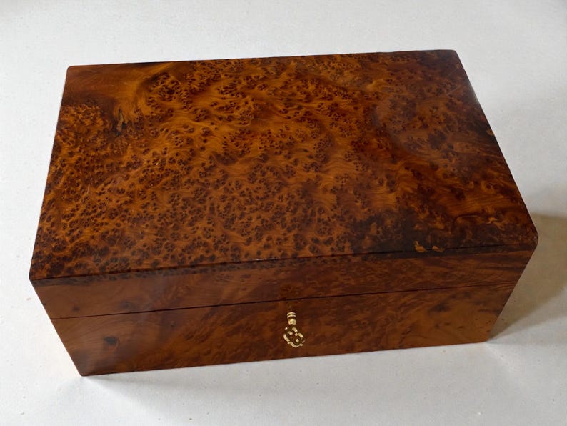 May include: A rectangular wooden box with a rich, warm brown burl wood grain finish. The box has a small, ornate gold-colored keyhole and a matching pull. The wood grain features a unique, swirling pattern.