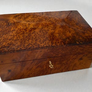 May include: A rectangular wooden box with a glossy finish. The box is a warm brown color with a unique burl wood grain pattern. It features a small, ornate gold-colored keyhole and appears to be a jewelry or keepsake box.