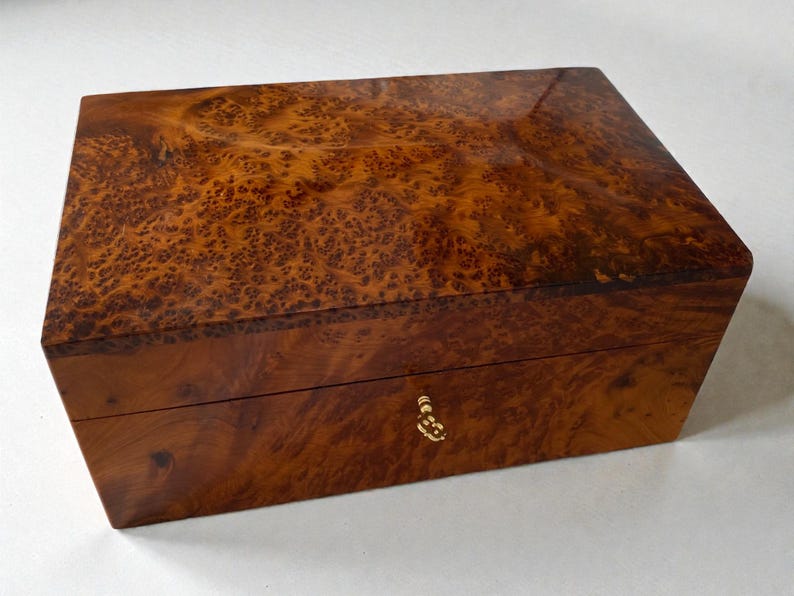 May include: A rectangular wooden box with a glossy finish. The box is a warm brown color with a unique burl wood grain pattern. It features a small, ornate gold-colored keyhole and appears to be a jewelry or keepsake box.