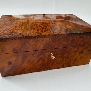 May include: A rectangular wooden box with a glossy finish. The box is made of a warm-toned wood with a unique burl grain pattern. It features a gold-colored clasp on the front. The box is likely used for storage or display.