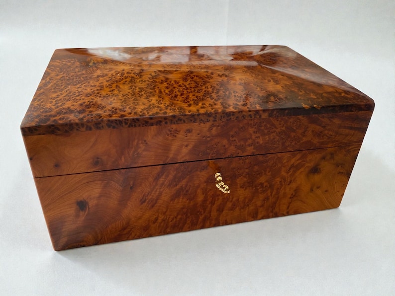 May include: A rectangular wooden box with a glossy finish. The box is made of a warm-toned wood with a unique burl grain pattern. It features a gold-colored clasp on the front. The box is likely used for storage or display.