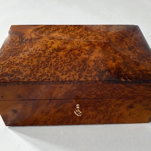 May include: A rectangular wooden box with a rich, warm brown burl wood grain finish. The box has a hinged lid and a small, ornate gold-colored keyhole. The wood grain features a unique pattern.