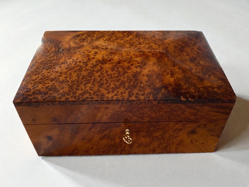 May include: A rectangular wooden box with a rich, warm brown burl wood grain finish. The box has a hinged lid and a small, ornate gold-colored keyhole. The wood grain features a unique pattern.