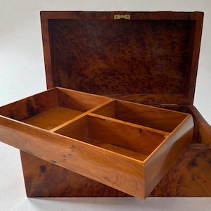 May include: Open wooden jewelry box with a removable tray. The box is made of a warm-toned wood with a burl grain pattern. The tray is divided into four compartments. The lid is open, revealing the interior.