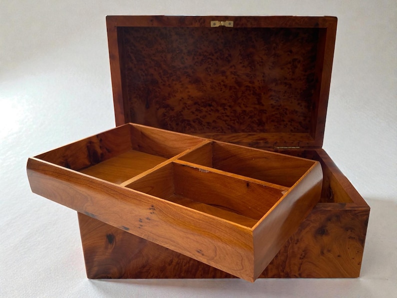 May include: Open wooden jewelry box with a removable tray. The box is made of a warm-toned wood with a burl grain pattern. The tray is divided into four compartments. The lid is open, revealing the interior.