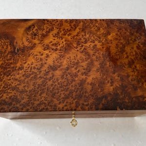 May include: A rectangular wooden box with a burl wood veneer. The box has a warm brown color with intricate patterns and a brass-colored keyhole and clasp. The box is likely used for storage.