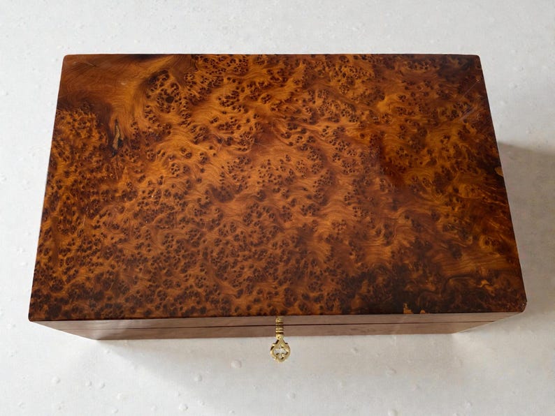 May include: A rectangular wooden box with a burl wood veneer. The box has a warm brown color with intricate patterns and a brass-colored keyhole and clasp. The box is likely used for storage.