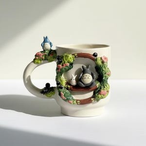 May include: Cream ceramic mug with a decorative design featuring characters from the animated film, My Neighbour Totoro. The handle and a frame around the mug are decorated with sculpted figures and floral accents. The mug has a glossy finish.