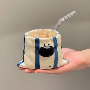 Könnte beinhalten: Ein cremefarbener Keramikbecher in Sackform mit vertikalen blauen Streifen. Der Becher hat eine markante schwarze ovale Nase und zwei kleine schwarze Punkte als Augen, die an ein Bärengesicht erinnern. Er ist mit Eiswürfeln und einem Glasstrohhalm.
