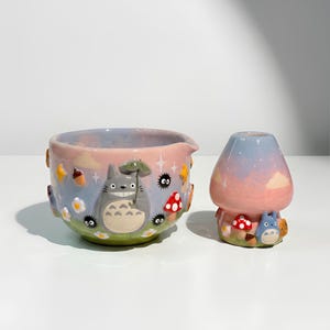 May include: A ceramic bowl and a matching mushroom-shaped container featuring a pastel sky background. The bowl displays a grey character holding a green umbrella, surrounded by small black soot sprites, red mushrooms, acorns, and white flowers.