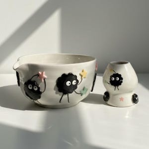 Puede incluir: Un juego de cuenco y jarrón de cerámica blanca. El cuenco tiene un pico y está decorado con personajes negros de dibujos animados que sostienen estrellas de colores. El jarrón tiene un diseño similar, con personajes negros y detalles de estrellas.