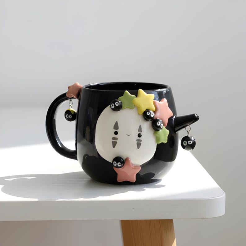 May include: A black ceramic mug featuring a white, oval-shaped face with two vertical gray markings. The mug is decorated with small, colorful pastel stars and several black soot sprite charms hanging from the handle and a small side protrusion.