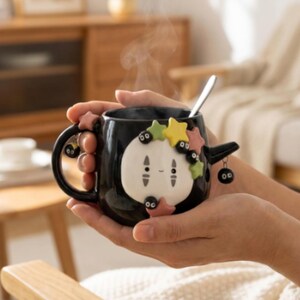 May include: A black ceramic mug featuring a white, oval-shaped character face with small black eyes and a subtle smile. The mug is decorated with colorful, raised star shapes and small black soot sprite figures. A silver spoon rests inside the mug.