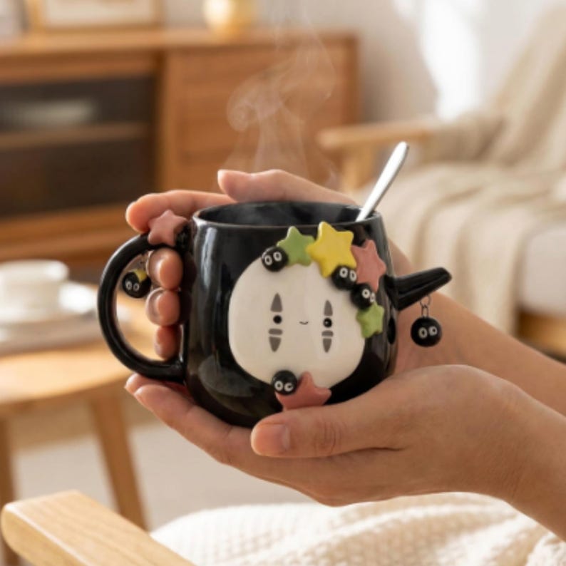 May include: A black ceramic mug featuring a white, oval-shaped character face with small black eyes and a subtle smile. The mug is decorated with colorful, raised star shapes and small black soot sprite figures. A silver spoon rests inside the mug.