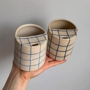 Könnte beinhalten: Zwei cremefarbene Wandtöpfe aus Keramik mit blauem Gittermuster. Jeder Topf hat eine abgerundete obere Öffnung und ein kleines Loch auf der Vorderseite. Die Töpfe werden in einer Hand gehalten, um ihre Größe und ihr Design zu zeigen. Geeignet für kleine Pflanzen oder Dekorationsartikel.