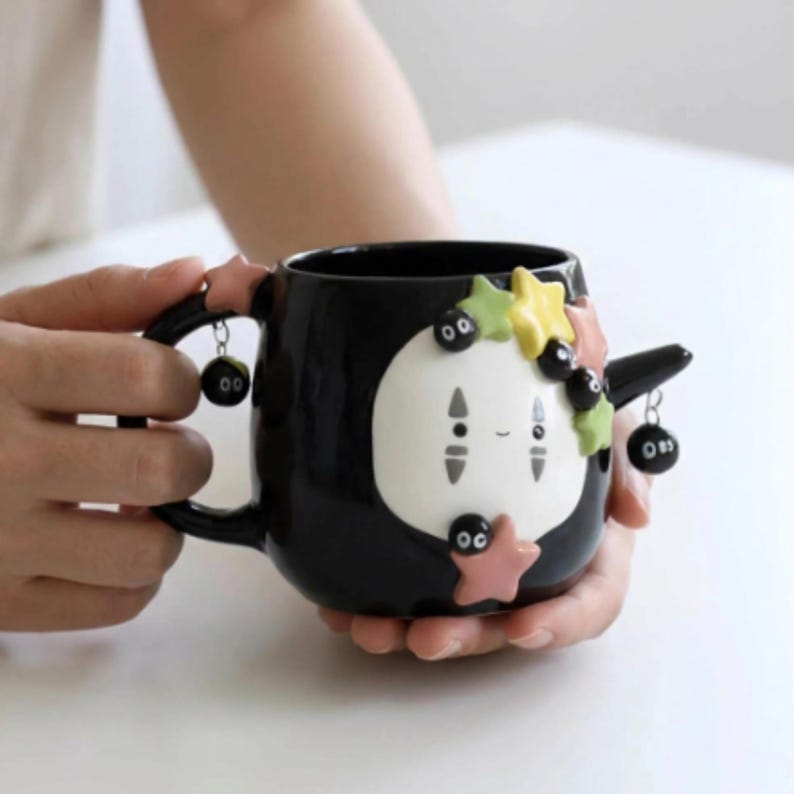 May include: A black ceramic mug featuring a white, oval-faced character with grey markings. The mug is decorated with small, colorful star-shaped accents and several dangling black soot sprite charms attached to the handle and a side spout.