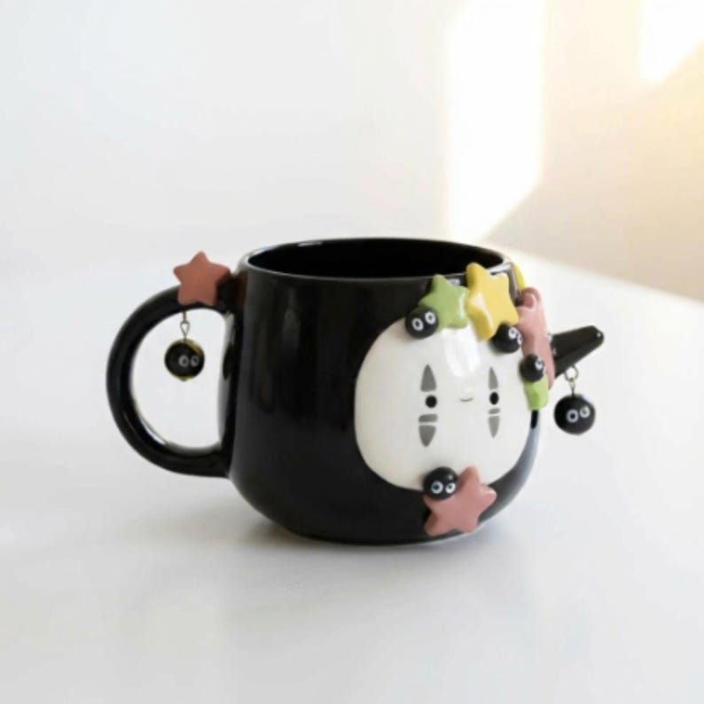 May include: A black ceramic mug featuring a white, mask-like face with simple facial features. The mug is decorated with small, colorful star-shaped appliques and dangling black soot sprite charms attached to the handle and side.