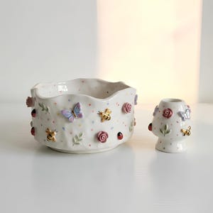 May include: A white ceramic bowl and a matching candle holder, both featuring a textured surface with colourful polka dots and three-dimensional embellishments including pink roses, blue butterflies, gold-accented bees, and red ladybirds.
