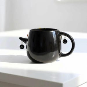 May include: A glossy black ceramic mug featuring a rounded body and a curved handle. A small, pointed protrusion extends from the side, and two miniature black spherical charms, each marked with the number 20, hang from silver-toned metal rings.
