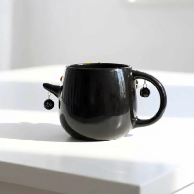 May include: A glossy black ceramic mug featuring a rounded body and a curved handle. A small, pointed protrusion extends from the side, and two miniature black spherical charms, each marked with the number 20, hang from silver-toned metal rings.