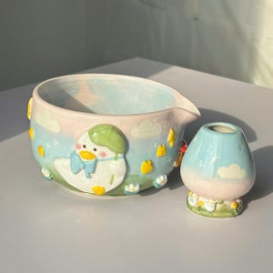 Puede incluir: Un cuenco vertedor de cerámica y un pequeño jarrón a juego con un fondo degradado en azul cielo y rosa pastel. El cuenco muestra una figura de pato blanco en relieve con sombrero verde y lazo azul, rodeado de flores amarillas y nubes blancas.
