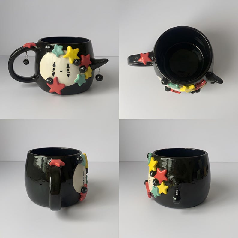 May include: A black ceramic mug featuring a white circular face with two vertical lines and a small mouth. The mug is decorated with raised yellow, red, and teal stars and small black soot sprites. A small black charm hangs from the handle.