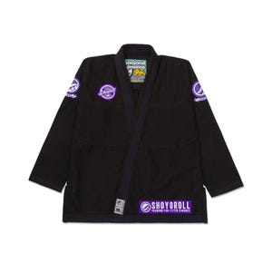 Könnte beinhalten: Schwarzer Jiu-Jitsu-Gi mit lila Besatz und gestickten Logos. Der Gi zeigt den Text "SHOYOROLL" in Weiß und Lila. Der Gi hat einen traditionellen Kragen und verstärkte Nähte. Der Gi ist für Kampfsporttraining und Wettkämpfe konzipiert.
