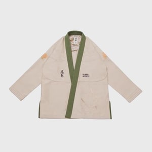 May include: A cream-coloured martial arts uniform with a green collar and trim. The uniform features Japanese characters and the text "ALBINO & PRETO". The sleeves and lapel have decorative patches.