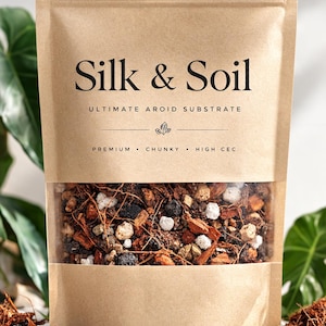 Puede incluir: Una bolsa de papel marrón con la inscripción "Silk & Soil" y una ventana transparente que revela una mezcla de sustrato aroide grueso. El sustrato contiene varios materiales, incluyendo trozos blancos, negros y marrones. La bolsa también tiene las palabras "Ultimate Aroid Substrate" y "Premium, Chunky, High CEC".