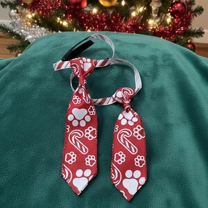 Pode incluir: Duas gravatas vermelhas e brancas para cães com tema natalino. As gravatas apresentam pegadas brancas e desenhos de bengalas doces. As gravatas estão sobre uma superfície verde, com uma árvore de Natal ao fundo.