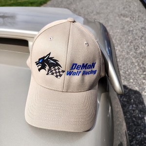 Peut inclure: Casquette de baseball beige avec une visière incurvée. Le chapeau présente un loup bleu et noir et le texte "DeMoN Wolf Racing" en bleu. La casquette est posée sur une voiture argentée.