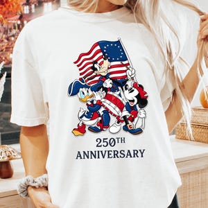May include: Cream-colored t-shirt featuring a graphic of Mickey Mouse, Donald Duck, and Minnie Mouse holding an American flag. The text "250th ANNIVERSARY" is below the characters. The shirt is paired with black shorts.