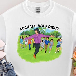 May include: White t-shirt with the text "MICHAEL WAS RIGHT" above an illustration of a person in a purple shirt and black pants, surrounded by running children. The shirt is paired with blue jeans and pink shoes.