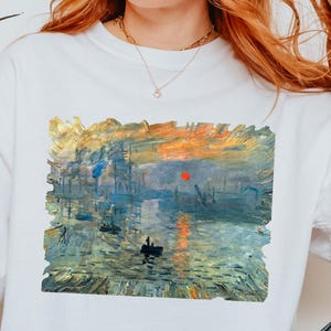 May include: White t-shirt featuring a colorful impressionist painting of a harbor scene with boats and a sunset. The artwork is framed within a rectangle on the front of the shirt. The shirt is paired with blue jeans.