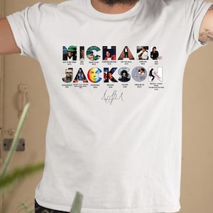 May include: A white short-sleeved t-shirt featuring a graphic print of the text MICHAEL JACKSON. The letters contain small images of album covers and music videos, with a signature printed below the text in the center of the chest area.